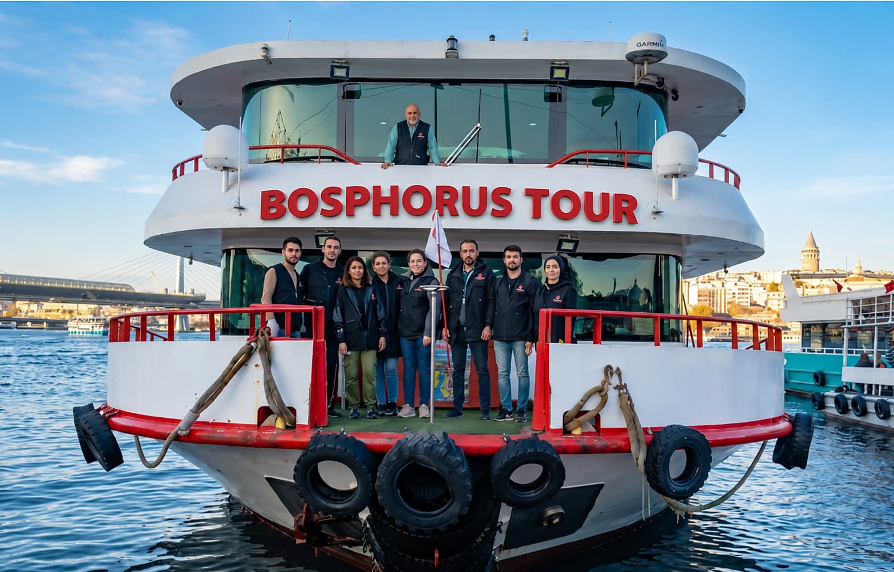 Crew standing on the bow of a Bosphorus tour boat in daylight, with Galata Tower in the background